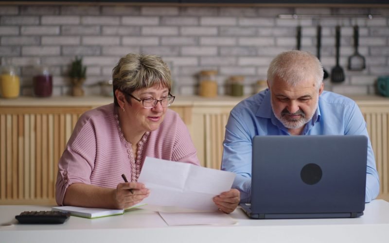 elderly-man-and-woman-are-filling-report-in-laptop-at-home-female-accountant-is-counti-SBI-351321907